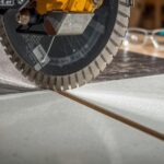 A close-up of a wet tile saw cutting a large-format porcelain tile cleanly, with water cooling the blade and safety glasses visible in the background.