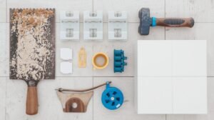 A complete set of tile installation tools laid out on a clean subfloor, including a notched trowel, tile spacers, rubber mallet, grout float, chalk line, and porcelain tiles.