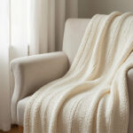 A cream knit throw draped over a linen armchair near a window with sheer curtains.