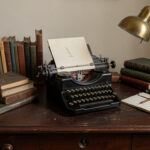 A dark wood writing desk with a vintage typewriter, stacked books, and a brass desk lamp.