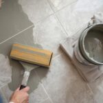 A homeowner applying grout to a newly tiled bathroom floor using a rubber float, with clean tile joints and a bucket of grout mixture visible nearby.