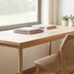 A light wood desk positioned beneath a large window with a woven chair and stacked notebooks.
