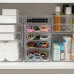 A neatly organized bathroom cabinet shelf with clear stackable acrylic bins holding cotton pads, hair ties, and small toiletries.
