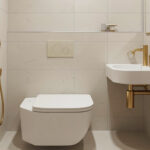 A slim wall-hung toilet and compact corner sink in a small luxury bathroom with light tile and brushed gold fixtures.