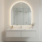 backlit arched LED mirror above a floating vanity, reflecting a frameless glass shower in a bright, minimalist bathroom.